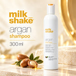 A 300 ml bottle of milk shake argan shampoo stands on a marble surface with argan nuts and leaves in the foreground, against a warm, softly lit background with gold-toned lighting and text on the left.