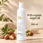 A white bottle of Milk Shake Argan Shampoo stands on a stone surface with argan nuts and leaves around it. Text on the right reads 'With organic argan oil' and '300 ml'.